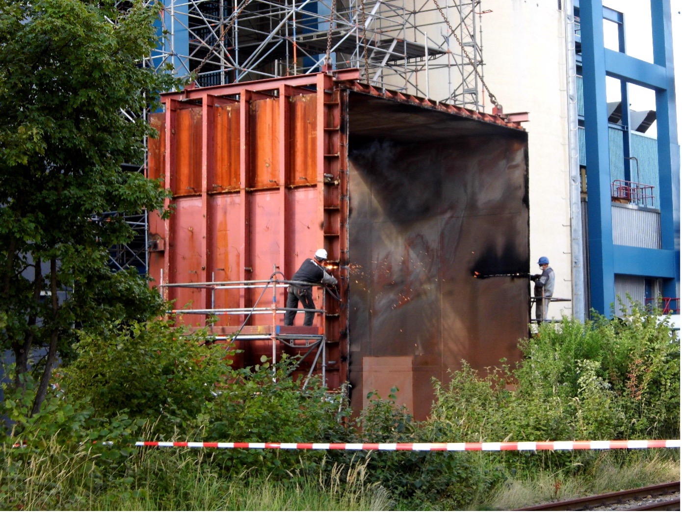 Zwei Arbeiter schweißen mit einem Gerät von Filtertechnik eine große Metallkonstruktion auf einer mit Pflanzen begrünten, mit Gerüsten versehenen Baustelle.