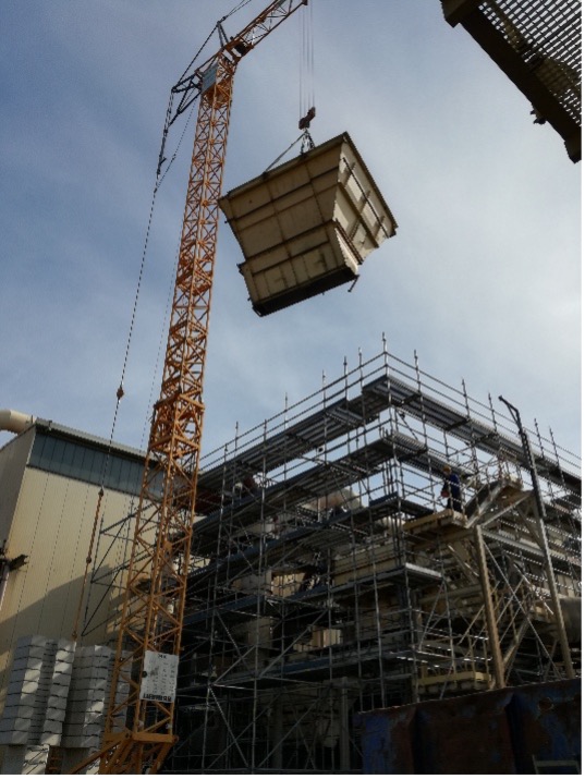 Ein Baukran hebt bei klarem Himmel einen Container mit Filtertechnik neben ein eingerüstetes Gebäude.