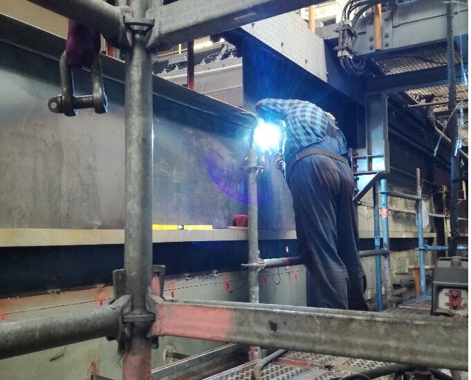 Person in Schutzkleidung schweißt Metallträger mit Filtertechnik auf Baustelle, umgeben von Gerüsten.