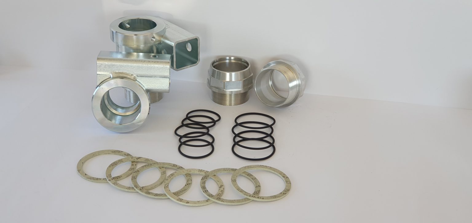 Verschiedene Metallverbinder, schwarze O-Ringe und flache Ersatzteil-Dichtungen liegen auf einer weißen Fläche vor weißem Hintergrund.